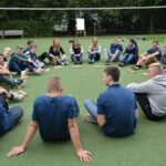 Gruppe Jugendlicher sitzt im Kreis auf Rasenplatz.