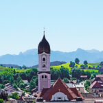 Dorf mit Kirchturm und Alpen im Hintergrund